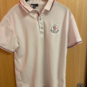 Vit pikétröja från Monclear - Säljer en stilren vit pikétröja från Hanyi Classic Fashion med Moncler-inspirerad logga på bröstet. Tröjan har korta ärmar och en klassisk krage med röd och svart randdetalj. Perfekt för en avslappnad men ändå snygg look.