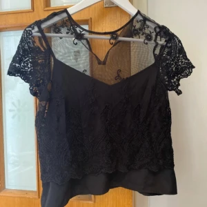 Svart spetsblus från Vero Moda - Säljer en elegant svart blus från Vero Moda med vackra spetsdetaljer och korta ärmar. Blusen har en transparent överdel och en underdel i ett mjukt tyg. Perfekt för en stilfull look. Tråden har gått av där bak för att sätta fast den men går att sy!