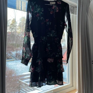 Svart blommig klänning från Ü - Säljer en svart klänning med blommigt mönster från Ü. Klänningen har långa ärmar och en volangdetalj vid kjolen.inga tecken på användning🩷