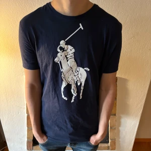 Mörkblå t-shirt från Polo Ralph Lauren - Säljer en mörkblå t-shirt från Polo Ralph Lauren med ett stort vitt polospel-tryck på framsidan. T-shirten är kortärmad och tillverkad i bomull, perfekt för en avslappnad stil.