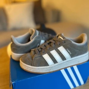 Grå Adidas Campus sneakers - Säljer ett par grå Adidas Campus sneakers med vita ränder och logga. Skorna har en klassisk design med snörning och är tillverkade i mocka. Perfekta för en avslappnad stil. De är dock lite slitna i sulan och har ett rivmärken men det märks ej. Storlek 35 och kommer med låda. Lådan är i acceptabelt skick men går ändå att använda.