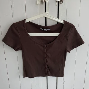 Brun ribbad kortärmad topp från 157 - Säljer en brun ribbad topp från 157 i storlek XS. Toppen är kortärmad och croppad med knappar framtill.