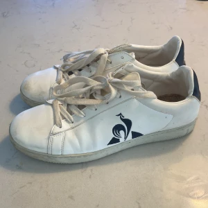 Vita sneakers från Le Coq Sportif - Säljer ett par vita sneakers från Le Coq Sportif med mörkblå detaljer och snörning. Skorna har en klassisk design med märkets logotyp på sidan. 
