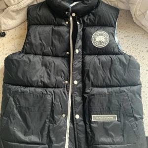 Svart dunväst från Canada Goose - Säljer en svart dunväst från Canada Goose med en snygg quiltad design. Västen har en dragkedja och knappar framtill samt praktiska fickor. Insidan har ett coolt mönster i blått och vitt. Perfekt för kyliga dagar!