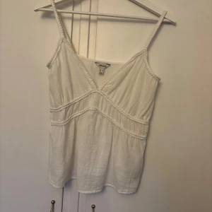Vit linnetopp  - Säljer en vit linnetopp med smala axelband och v-ringad design. Toppen har dekorativa sömmar och en luftig passform, perfekt för varma dagar.