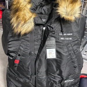 Svart parkajacka från Alpha Industries - Säljer en svart parkajacka från Alpha Industries med pälsfodrad huva och dragkedja. Jackan har flera fickor och detaljer som knappar och broderad text. Perfekt för kalla dagar med sin robusta design och varma foder.