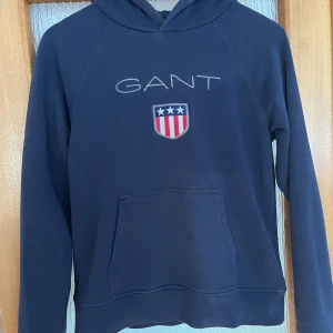 Mörkblå hoodie från GANT - Säljer en snygg mörkblå hoodie från GANT med broderad logga och emblem på bröstet. Tröjan har en klassisk känguruficka och långa ärmar. Perfekt för en avslappnad stil.