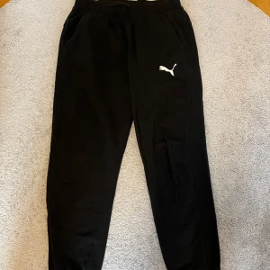 Svarta mjukisbyxor från Puma - Säljer ett par svarta mjukisbyxor från Puma med elastisk midja och det klassiska Puma-logotypen på benet. Perfekta för en avslappnad stil eller träning.