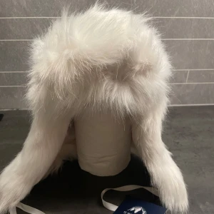 Vit pälsmössa från Newport - Säljer en lyxig vit pälsmössa från Newport, St. Moritz Faux Fur White Fox Hat. Mössan är i storlek small/medium och har ett mjukt, fluffigt utseende med knytband under hakan. Perfekt för kalla vinterdagar. Helt ny🥰nypris 1500kr