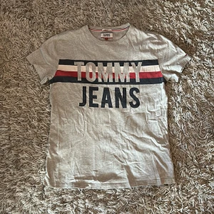 Grå t-shirt från Tommy Jeans - Säljer en grå t-shirt från Tommy Jeans med stort tryck i mörkblå, rött och vitt på framsidan. T-shirten har en klassisk passform och är kortärmad. Perfekt för en avslappnad stil.