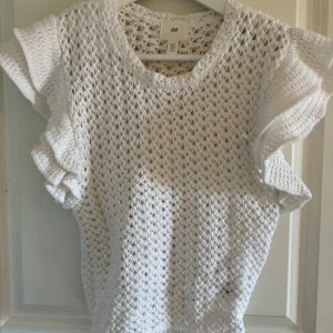 Vit stickad topp från H&M - Säljer en vit stickad topp från H&M med ett luftigt mönster. Toppen har korta ärmar med volangdetaljer som ger en söt touch. Perfekt för varmare dagar eller som ett lager över en annan topp.