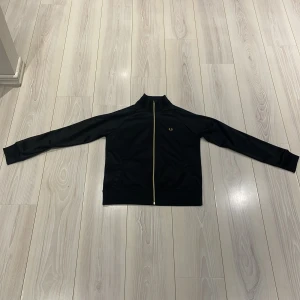 Svart tröja med zip från Fred Perry - Säljer en stilren svart zip up tröja från Fred Perry med gulddragkedja och broderad logga på bröstet. Jackan har långa ärmar och en klassisk passform. Nypris ligger på 1500kr, Perfekt för en sportig look eller casual vardagsstil.