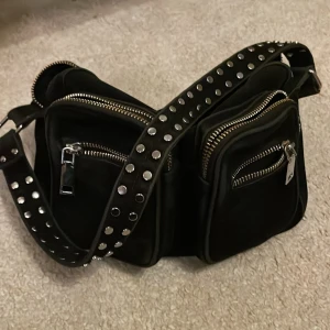 Svart axelväska med nitar - Äkta noella! Snygg svart axelväska med flera dragkedjefickor och coola nitar på axelremmen. Perfekt för att hålla ordning på dina grejer med stil. Väskan har en rymlig insida och flera praktiska fack. Man ser på bilderna att färgen gått bort lite men inget som syns på långt håll!