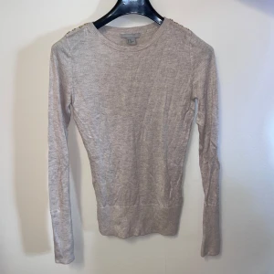 Beige tröja från H&M - Säljer en stilren beige tröja från H&M i storlek XS. Tröjan har långa ärmar och en rund halsringning. Har gulddetaljer på axlarna.  Perfekt för en enkel och elegant look. Passar bra till både jeans och kjol.