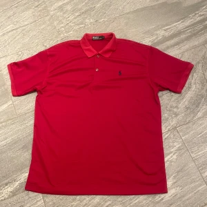 Röd pikétröja från Polo Ralph Lauren - Snygg röd pikétröja från Polo Ralph Lauren med klassisk krage och knappslå. Den har en broderad logga på bröstet i blått. Perfekt för en stilren look. Passar ungefär som en storlek L-XL