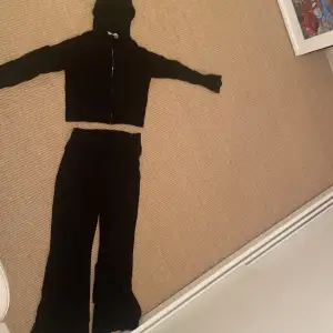 Säljer en svart ribbad hoodie med dragkedja och matchande byxor. Hoodien har en huva och långa ärmar, perfekt för en avslappnad stil. Byxorna är i samma ribbade material och ger en enhetlig look.Tröjan är gjord som en magtröja och därför är lite kortare byxorna är också bootcut 