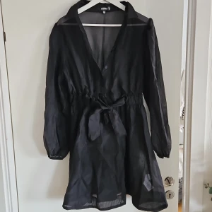 Svart klänning från Missguided - Snygg svart klänning från Missguided med knytband i midjan och långa ärmar. Den har en transparent look och en v-ringad design som ger en elegant touch. Perfekt för att skapa en stilren outfit.