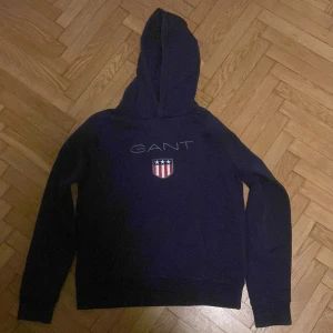Mörkblå hoodie från GANT - Säljer en snygg mörkblå hoodie från GANT med deras logga och ett emblem på bröstet. Tröjan har långa ärmar och en klassisk huva. Plagget är i barnstorlek.
