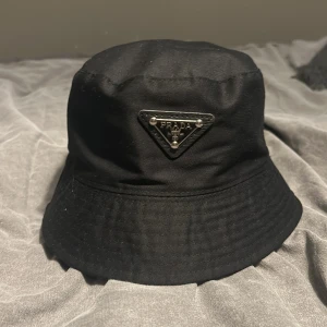 Svart bucket hat från Prada - Snygg svart bucket hat från Prada med den ikoniska triangulära logotypen framtill. Perfekt för att ge din outfit en stilren touch. Tillverkad i ett slitstarkt material som passar bra för olika säsonger.
