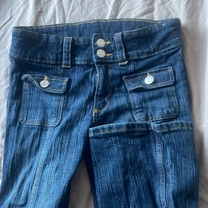 bootcut jeans från H&M - Snygga blå bootcut jeans från H&M med låg midja och justerbar midja. De har dekorativa knappar och fickor fram. Perfekta för en avslappnad stil.