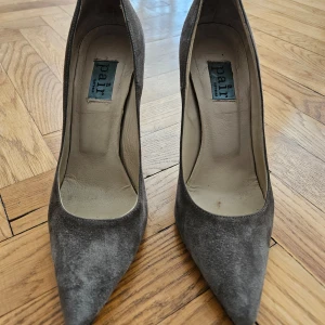 Grå mockapumps från "a Pair" - Snygga grå mockapumps från danska  a Pair med spetsig tå och 10 cm klack. Mocka, tå och klack är helt oskadade. På sula syns vanlig "wear and tear". Nypris SEK 3000