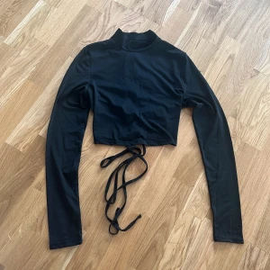 Svart långärmad croppad topp - Snygg svart croppad topp med långa ärmar och hög krage. Toppen har en unik snörning i ryggen som ger en cool detalj. Perfekt för att skapa en stilren look.