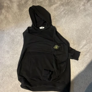 Svart hoodie med emblem - Snygg svart hoodie med en klassisk design och ett emblem på ärmen. Perfekt för en avslappnad stil. Den har en stor ficka framtill och en bekväm huva.