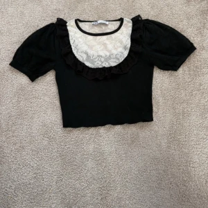 Svart topp med spetsdetaljer - Snygg svart topp från Zara med korta puffärmar och en vacker spetsdetalj framtill. Toppen har en volangkant runt halsen som ger en romantisk touch. Perfekt för att piffa upp din outfit!