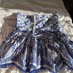 Vit och blå kjol från Zara - Säljer en superfin vit och blå kjol från Zara med ett detaljerat blommönster. Kjolen har knappar framtill och en volangdetalj. Perfekt för en somrig look, 