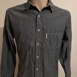 Mörkblå skjorta från Ruffle & Bow - Snygg mörkblå skjorta från Ruffle & Bow med lång ärm och klassisk krage. Skjortan har en bröstficka och små broderade detaljer. Perfekt för en stilren look.