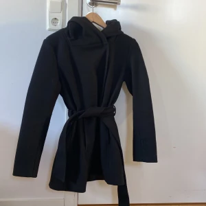Svart kappa från New Yorker  - Snygg svart kappa från New Yorker med huva och knytbälte i midjan. Den har en stilren design med knappar framtill och långa ärmar. Perfekt för kyligare dagar.