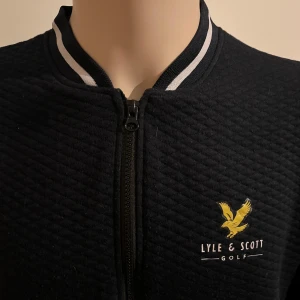 Svart quiltad tröja från Lyle & Scott - Snygg svart quiltad tröja från Lyle & Scott med dragkedja framtill. Jackan har en broderad logga på bröstet och ribbade detaljer vid kragen. Perfekt för golf eller andra aktiviteter. Sprättade bort lappen,den kliade
