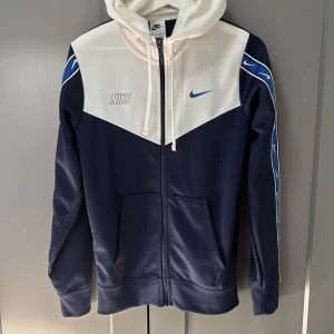 Blå och vit hoodie från Nike - Snygg hoodie från Nike i blått och vitt med dragkedja och huva. Den har långa ärmar med coola mönster längs sidorna och Nike-logga på bröstet. Perfekt för en sportig look!
