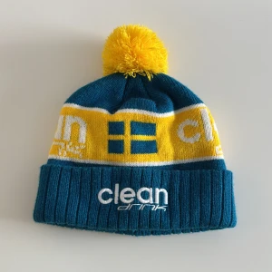 Clean drink mössa - Snygg mössa i blått och gult med en stor gul tofs på toppen. Mössan har ett broderat märke 'clean drink' och ett svenskt flaggmönster. Perfekt för kalla dagar.