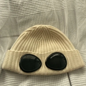 Beige ribbad mössa från C.P. Company - Snygg grädd vit  ribbad mössa från C.P. Company med unika svarta glasögondetaljer framtill. Du får även med ett armband Cordell 