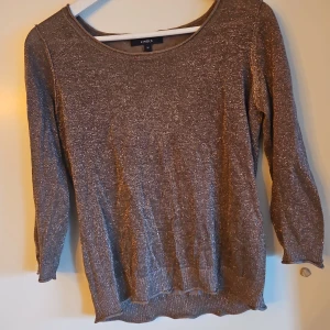 Glittrig tröja från Lindex - Snygg glittrig tröja från Lindex i en skimrande brun nyans. Tröjan har trekvartsärmar och en rund halsringning, perfekt för att ge lite extra glans till din outfit. Passar bra till både vardag och festliga tillfällen.