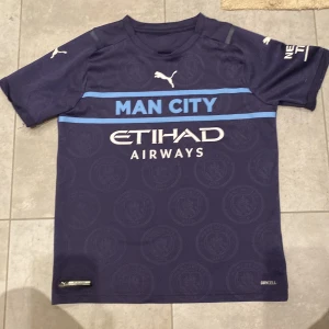 Mörkblå Man City fotbollströja från Puma - Snygg mörkblå Man City fotbollströja från Puma med korta ärmar. Tröjan har ljusblå detaljer och vit text med 'Etihad Airways' tryckt på framsidan. Perfekt för alla Man City-fans!