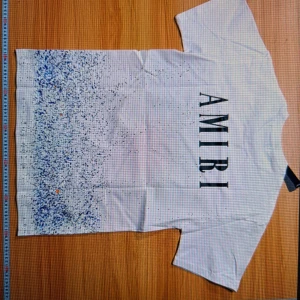 Vit t-shirt från Amiri med blått stänkmönster - Säljer en vit t-shirt från Amiri med ett coolt blått stänkmönster nertill. T-shirten har korta ärmar och ett stort Amiri-tryck på framsidan. Perfekt för en avslappnad stil.