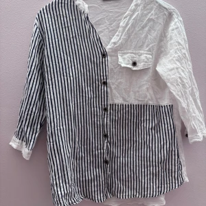 Randig och vit skjorta med knappar - Unik skjorta med en mix av randigt och vitt tyg. Ena sidan och ena ärmen är randiga i svart och vitt, medan resten är helvit. Skjortan har knappar framtill och en bröstficka. Perfekt för en stilren look.