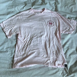 Ljusrosa t-shirt från Pull&Bear - Säljer en ljusrosa t-shirt från Pull&Bear med tryck på både fram- och baksida. På baksidan står det 'Malibu Bay Paradise Place' med en krabba och texten 'California'. Perfekt för en avslappnad stil. 