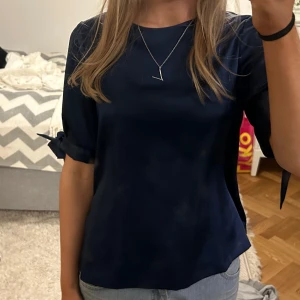 Marinblå satin blus bymalina - Säljer en stilren marinblå satin blus med korta ärmar med rosetter. Perfekt för en elegant och avslappnad look. Blusen har en mjuk och skön passform.