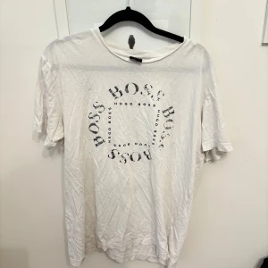 Vit t-shirt från Hugo Boss - Säljer en vit t-shirt från Hugo Boss med ett cirkulärt tryck av märkesnamnet på framsidan. T-shirten är kortärmad och har en avslappnad passform. .