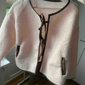 Vit teddyjacka med knytband - Säljer en mysig vit teddyjacka med brun detaljer och knytband framtill. Jackan har en rund hals och långa ärmar, perfekt för kyliga dagar. Den har också praktiska fickor med brun kanter.