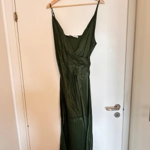 Grön långklänning med omlott - Zara - Snygg grön långklänning med omlott-design och smala axelband. Perfekt för en elegant look. Klänningen har en v-ringad fram och är tillverkad i ett glansigt material som ger en lyxig känsla. Tyvärr skrynklig då den inte använts på länge.