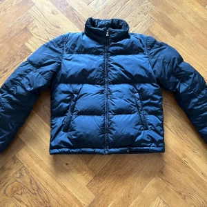Mörkblå dunjacka från Prada - Snygg svart dunjacka från Prada. AW09, red stripe logo. Perfekt skick och mycket hög kvalitet. Storlek 50 M/L croppad fit. Riktig grail ✨