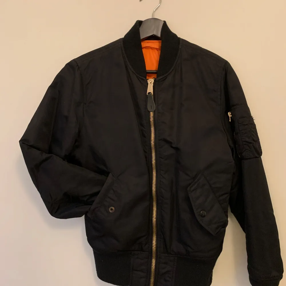 Säljer en klassisk svart bomberjacka från Alpha Industries. Jackan har en dragkedja framtill och är långärmad med ribbade muddar. Den har en orange insida och praktiska fickor med knappar. Perfekt för en cool och avslappnad stil. Lite nopprig på ryggen, se sista bilden. I övrigt jättefint skick!. Takit.