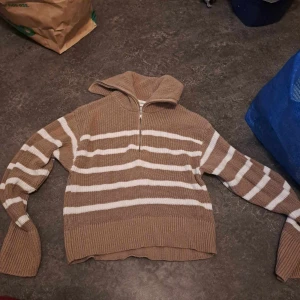 Brun och vit randig stickad tröja - Säljer en mysig brun och vit randig stickad tröja med dragkedja vid halsen. Tröjan har långa ärmar och en bekväm passform, perfekt för kyligare dagar.