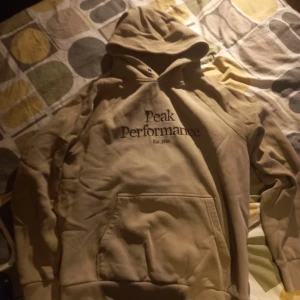 Beige hoodie från Peak Performance - Säljer en beige hoodie från Peak Performance med tryck på bröstet. Den har en klassisk känguruficka och en justerbar huva. Perfekt för en avslappnad stil.