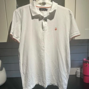 Vit pikétröja från Sail Racing - Säljer en stilren vit pikétröja från Sail Racing. Tröjan har korta ärmar och en klassisk krage med knappar. Detaljer i form av en liten logotyp på bröstet och orange kant vid ärmslut och krage. Perfekt för en avslappnad och sportig look.