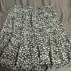 Blommig kjol från Menglu - Charmig blommig kjol från Menglu med elastisk midja. Kjolen har ett sött blommönster i vitt och grönt på en mörk bakgrund. Kjolen har också en volang där nere  Perfekt för en somrig look!